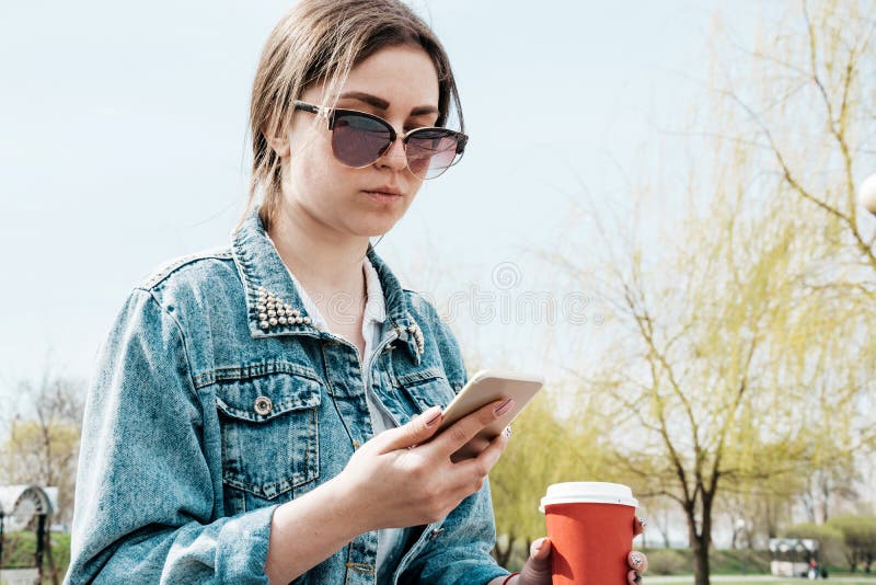 A young girl in a denim jacket walks down the street with coffee and a phone in her hands. Girl in glasses and a denim jacket on stock photo A young girl in a denim jacket walks down the street with coffee and a phone in her hands. Girl in glasses and a denim jacket on stock photo