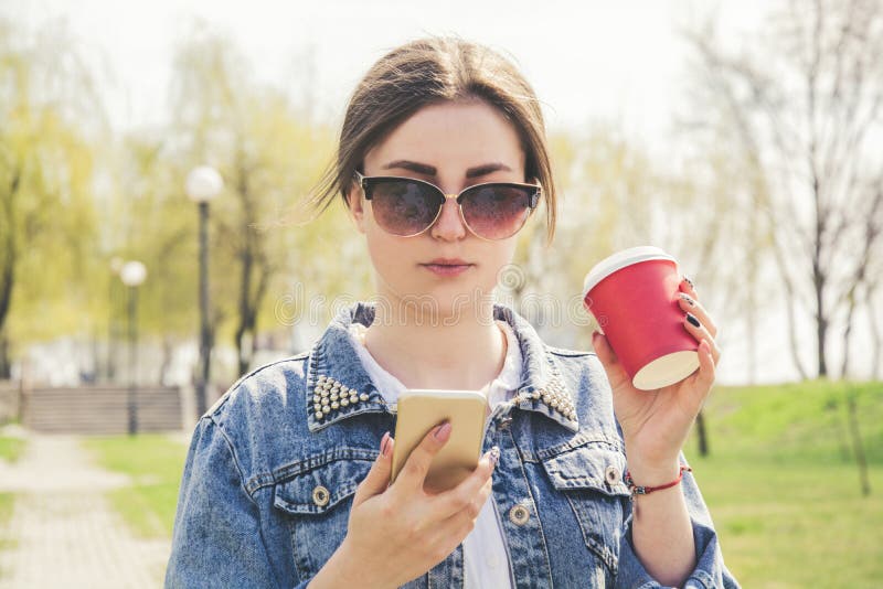 A young girl in a denim jacket walks down the street with coffee and a phone in her hands. Girl in glasses and a denim jacket on royalty free stock photography A young girl in a denim jacket walks down the street with coffee and a phone in her hands. Girl in glasses and a denim jacket on royalty free stock photography