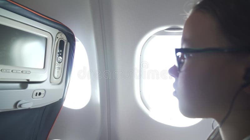 Young girl with glasses and headphones watches video on the monitor built into armchair in the cabin of the airplane