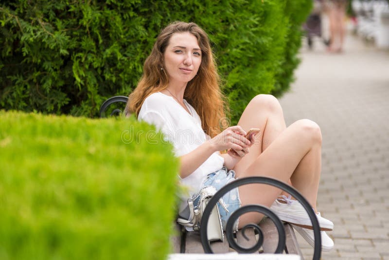 A young girl with phone in her hands, sits on a bench in the park and looked at the frame