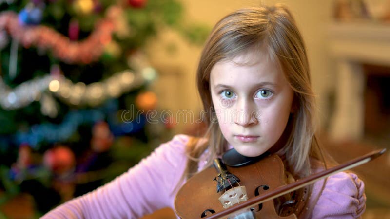 Young girl playing violin in front of christmas tree. Blonde young female musician violinist plays violin with Christmas