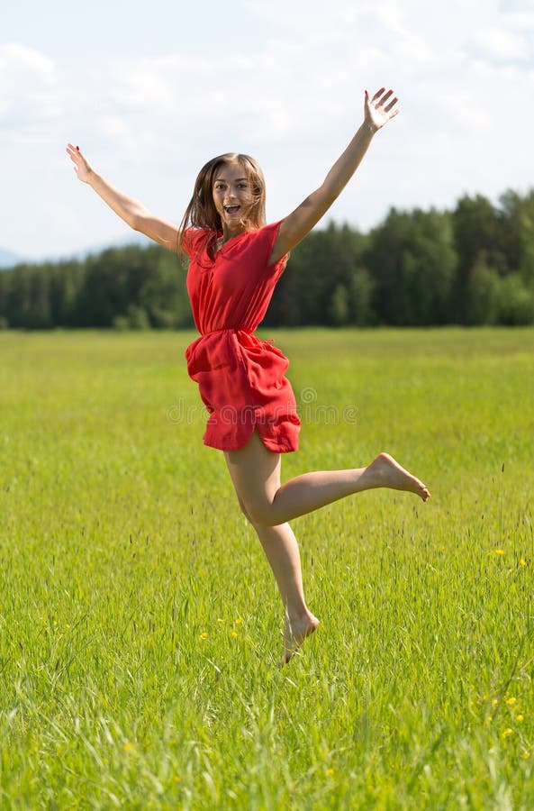 Young girl in a red dress jumping in a field with forest