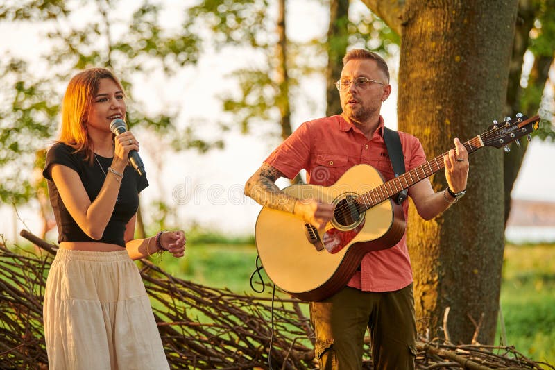 Young girl singer holds microphone in hand with man guitarist plays rock music at outdoor festival