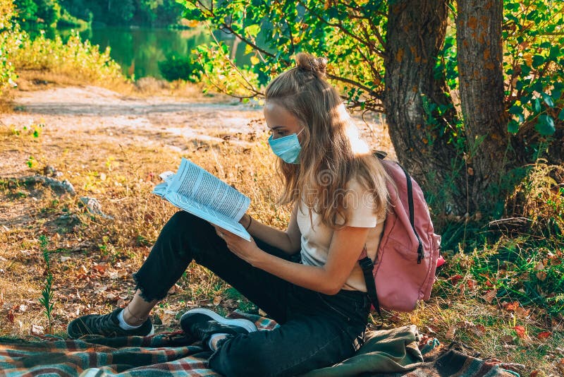 Young girl, student sitting outdoor with backpack and reading a book. Wearing protective face mask. Study from home. Cottagecore stock images Young girl, student sitting outdoor with backpack and reading a book. Wearing protective face mask. Study from home. Cottagecore stock images
