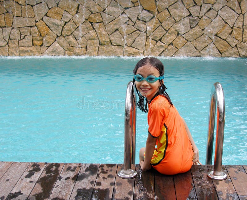 Little girl having fun at the pool - A picture of a young asian girl swimmer sitting on edge of a blue swimming pool with wooden deck and waterfall feature as background. Girl is in bright orange and yellow swim suit and smiling happily into camera. Model is 8 to 9 years old. Horizontal color format. Little girl having fun at the pool - A picture of a young asian girl swimmer sitting on edge of a blue swimming pool with wooden deck and waterfall feature as background. Girl is in bright orange and yellow swim suit and smiling happily into camera. Model is 8 to 9 years old. Horizontal color format.