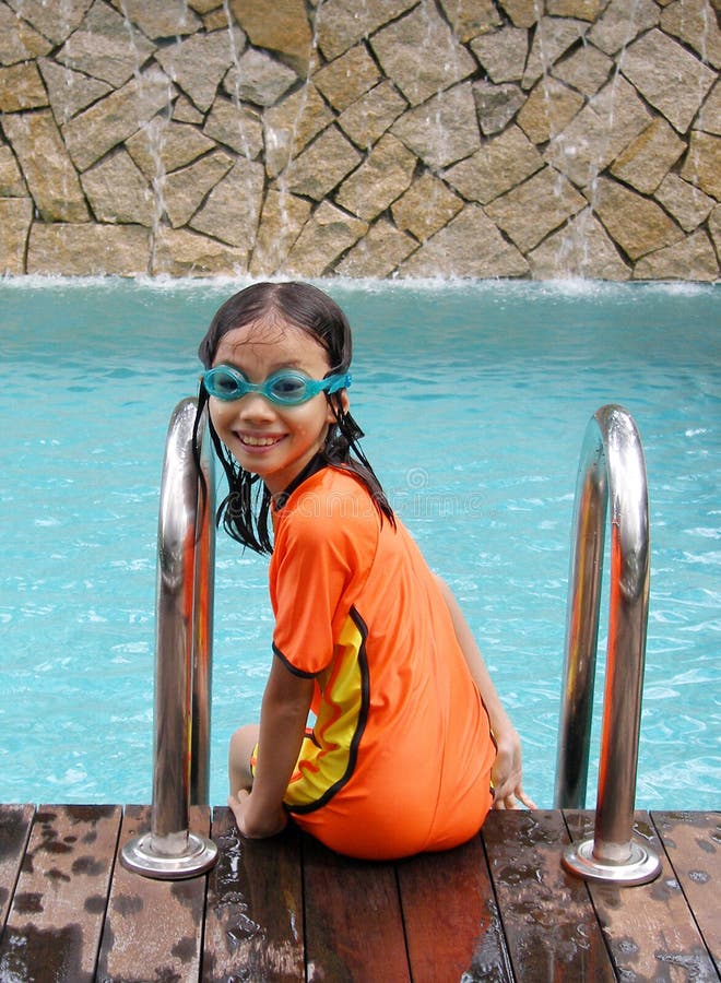 Kid having fun in the pool - A picture of a young asian girl swimmer sitting on edge of a blue swimming pool with wooden deck and waterfall feature as background. Little girl is in bright orange and yellow swim suit and smiling happily into camera. Model is 8 to 9 years old. Vertical format. Kid having fun in the pool - A picture of a young asian girl swimmer sitting on edge of a blue swimming pool with wooden deck and waterfall feature as background. Little girl is in bright orange and yellow swim suit and smiling happily into camera. Model is 8 to 9 years old. Vertical format.