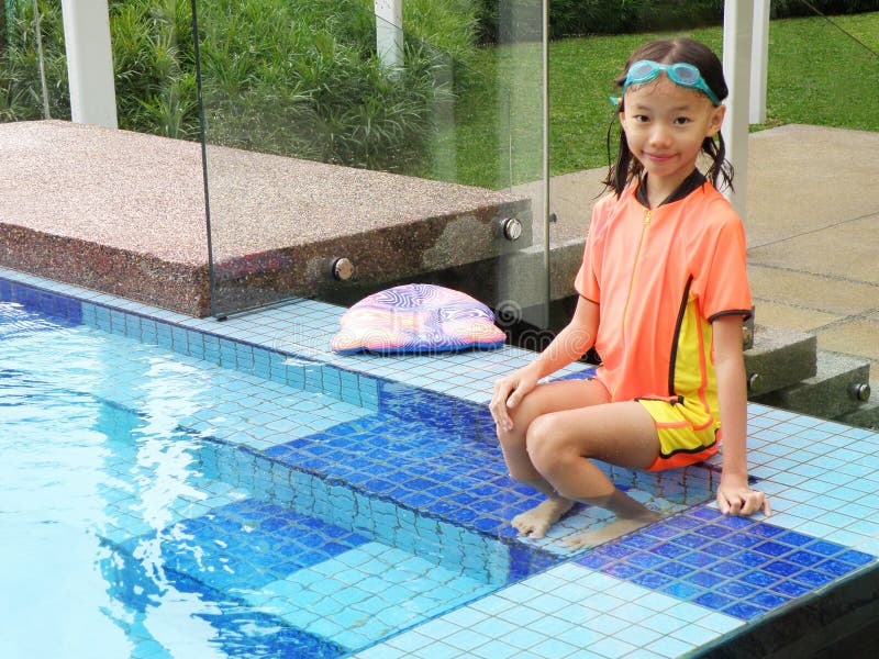 A picture of a young asian girl swimmer sitting on edge of a blue swimming pool with blue mosaic tiles. Little girl is in bright orange and yellow swim suit and smiling happily into camera. Model is 8 to 9 years old. Vertical color format. A picture of a young asian girl swimmer sitting on edge of a blue swimming pool with blue mosaic tiles. Little girl is in bright orange and yellow swim suit and smiling happily into camera. Model is 8 to 9 years old. Vertical color format.