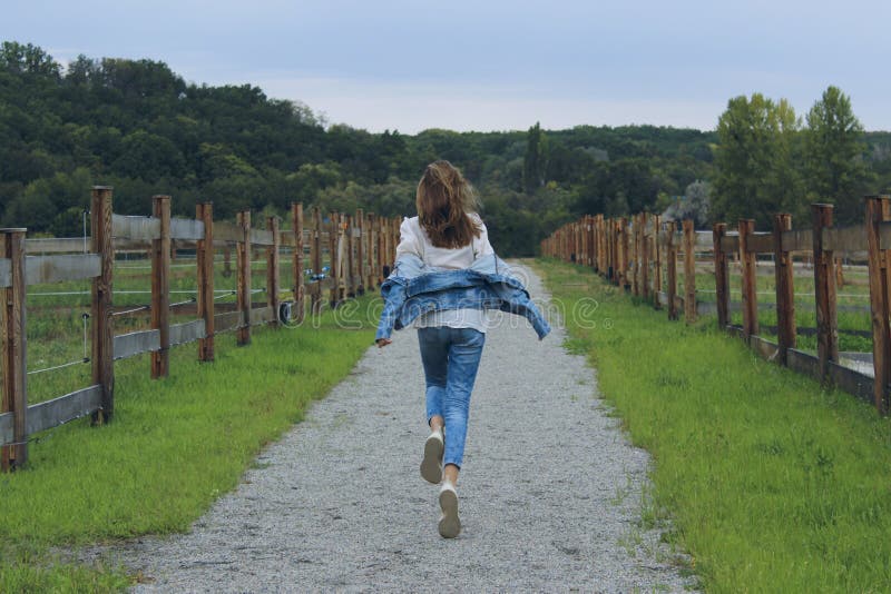 Young girl wearing a blue jeans and jeans jacket running outdoor. Girl running along the road in the stable. stock photo Young girl wearing a blue jeans and jeans jacket running outdoor. Girl running along the road in the stable. stock photo