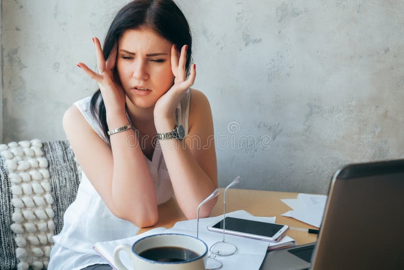 A young girl in white clothes holding her head on a gray background. Work in a cafe. On the desktop is coffee, computer, work royalty free stock image A young girl in white clothes holding her head on a gray background. Work in a cafe. On the desktop is coffee, computer, work royalty free stock image