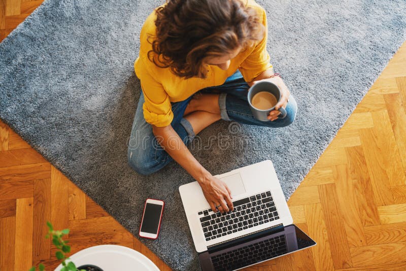 Young girl woman in a yellow shirt works on a laptop with a mug of coffee at home in the living room on the carpet, remote work
