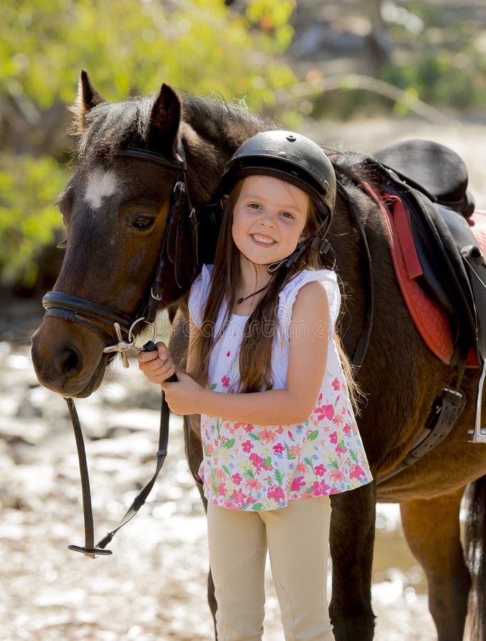 Young girl 7 or 8 years old holding bridle of little pony horse smiling happy wearing safety jockey helmet in summer holiday