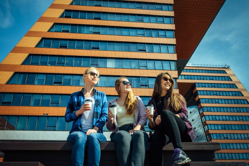 Young girls in sunglasses sitting on a city street and drink coffee to take away. Three cute women have a coffee break together stock image Young girls in sunglasses sitting on a city street and drink coffee to take away. Three cute women have a coffee break together stock image