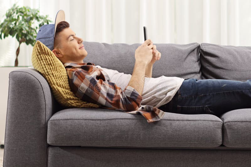 Man Looking His Smart Phone while, Lying on the Couch at Home Stock