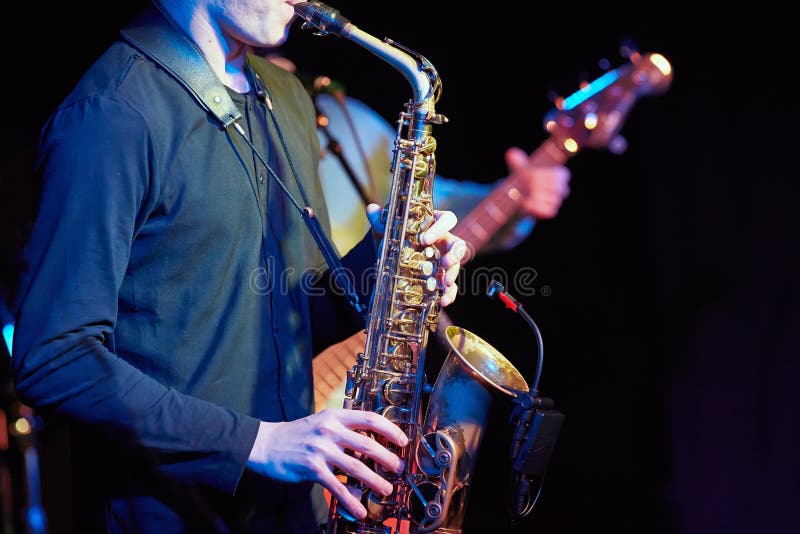 A young guy plays a saxophone with a microphone on the background of a guitarist on a dark background.