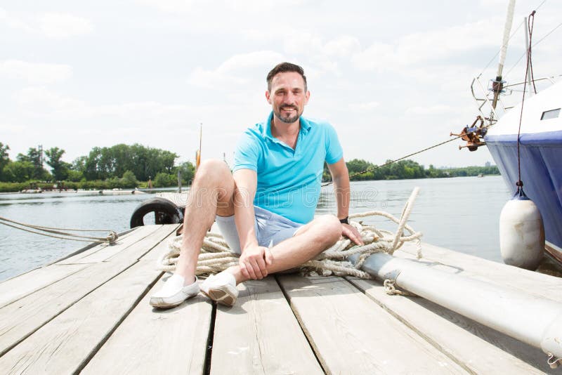 A young guy sits in blue polo on pier near the yacht. happy smiled tourist handsome man relaxing and enjoying the view on river. stock photography A young guy sits in blue polo on pier near the yacht. happy smiled tourist handsome man relaxing and enjoying the view on river. stock photography
