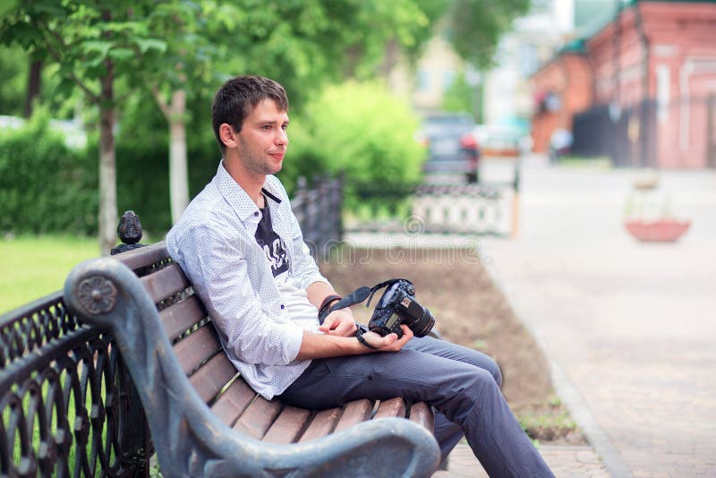 The young guy in a white shirt sits on a shop on the street in the park, holds the mirror camera in hand stock photo The young guy in a white shirt sits on a shop on the street in the park, holds the mirror camera in hand stock photo