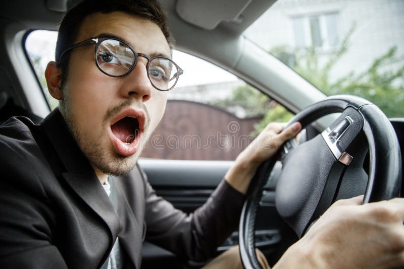 Young guy yawns while looking at the camera. He is sitting at his car. His hands are on the steering wheel