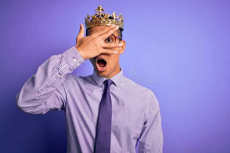 Young handsome african american man wearing golden crown of king over purple background peeking in shock covering face and eyes stock photos