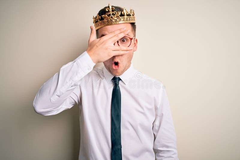Young handsome caucasian business man wearing golden crown over isolated background peeking in shock covering face and eyes with stock photography
