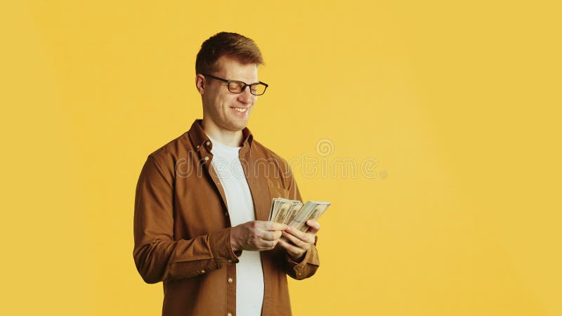 Young handsome caucasian man in glasses and casual clothes standing and counting the stack of banknotes, smiling