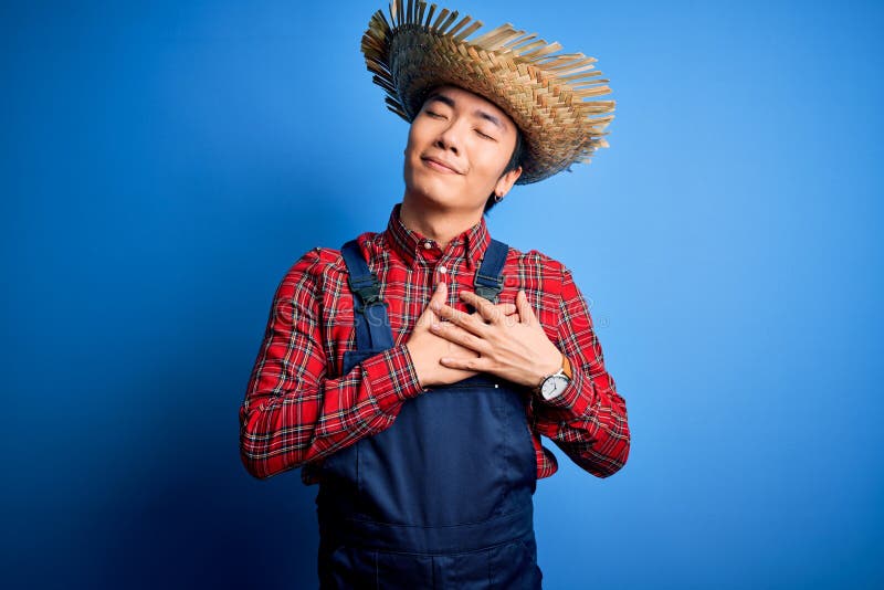 Young handsome chinese farmer man wearing apron and straw hat over blue background smiling with hands on chest with closed eyes