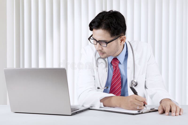 Doctor looking at his laptop while writing a prescription stock photos Doctor looking at his laptop while writing a prescription stock photos