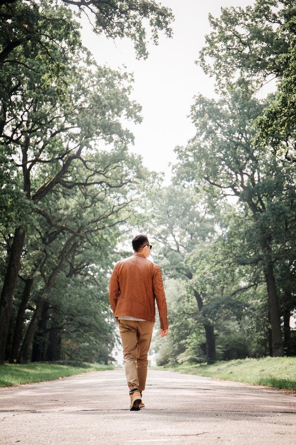 A Young Handsome Guy is Walking Along a Wide Road with Huge Trees, Back