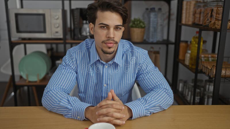 Young handsome hispanic man in office sitting confidently at desk with modern interior creating a professional work atmosphere in stock photography