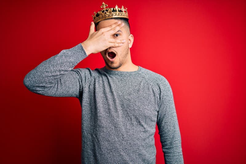 Young handsome man wearing golden crown of prince over isolated red background peeking in shock covering face and eyes with hand stock image