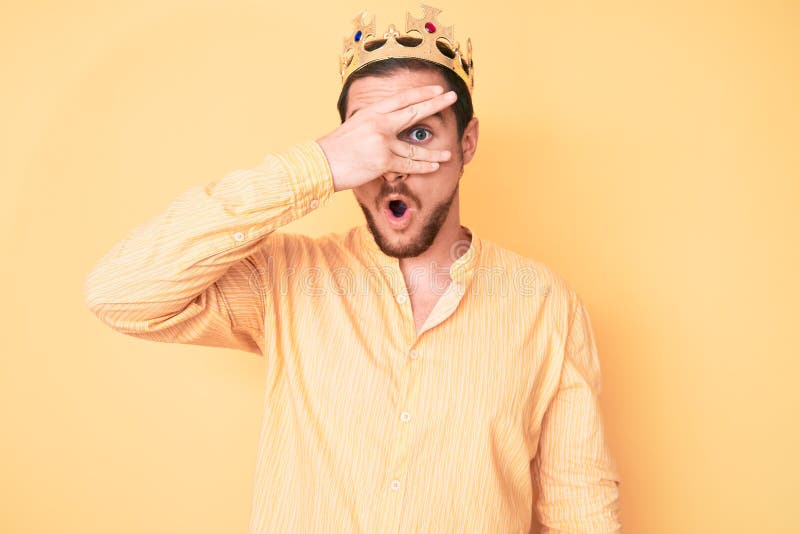 Young handsome man wearing king crown peeking in shock covering face and eyes with hand, looking through fingers afraid royalty free stock image