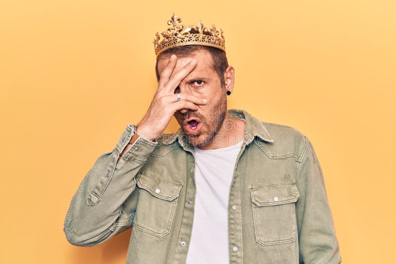 Young handsome man wearing king crown peeking in shock covering face and eyes with hand, looking through fingers afraid royalty free stock photo