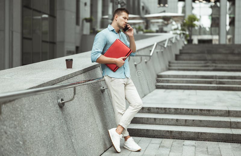 Young handsome office worker talking on the phone holding laptop standing on the stairs in the street during lunch break