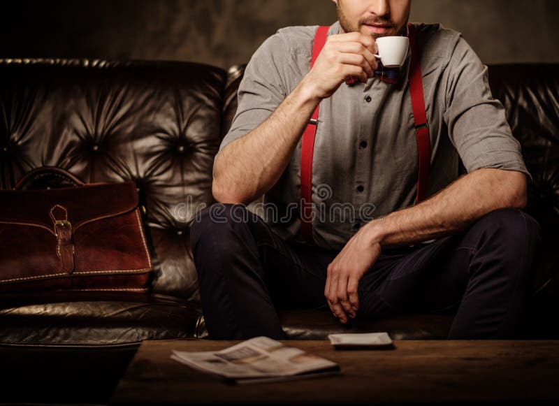 Young handsome old-fashioned bearded man with cup of coffee sitting on comfortable leather sofa on dark background. stock photography