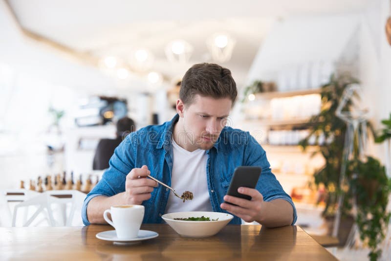 Young handsome smiling man drinks his hot coffee and eats salad for lunch while typing message in his smartphone during break at