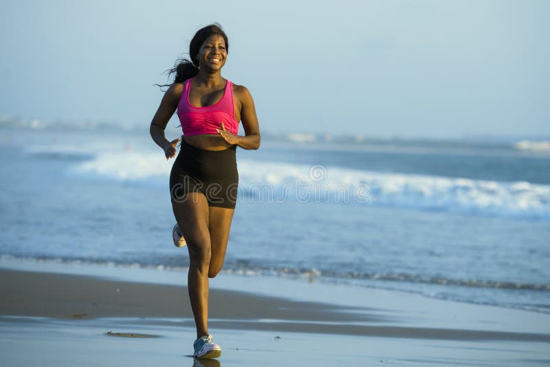 Young happy and attractive African American runner woman exercising on running workout at beautiful beach jogging and enjoying sun