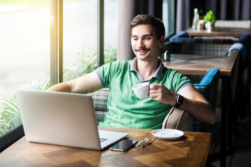 Young happy businessman in green t-shirt sitting and looking ot laptop display with smile, holding and drinking cup of coffee, royalty free stock image Young happy businessman in green t-shirt sitting and looking ot laptop display with smile, holding and drinking cup of coffee, royalty free stock image
