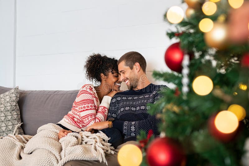 Young happy couple embracing and relaxing on comfortable couch. Lovely multiethnic couple sitting on sofa wearing christmas sweater and hugging. African woman and affectionate man near christmas tree. Young happy couple embracing and relaxing on comfortable couch. Lovely multiethnic couple sitting on sofa wearing christmas sweater and hugging. African woman and affectionate man near christmas tree