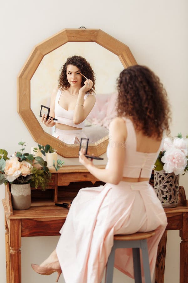 Young happy girl with curly hair does makeup in front of a vintage mirror. A beautiful woman in a pink dress paints her eyes