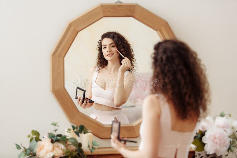 Young happy girl with curly hair does makeup in front of a vintage mirror. A beautiful woman in a pink dress paints her eyes
