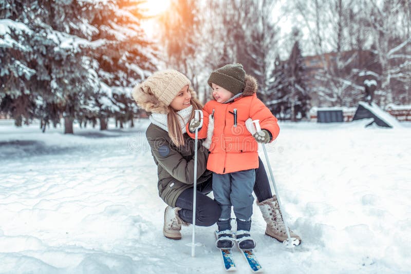 Young happy mother, a woman holds a boy 2-4 years old son, learns to ski. In winter in park outside. Free space for text