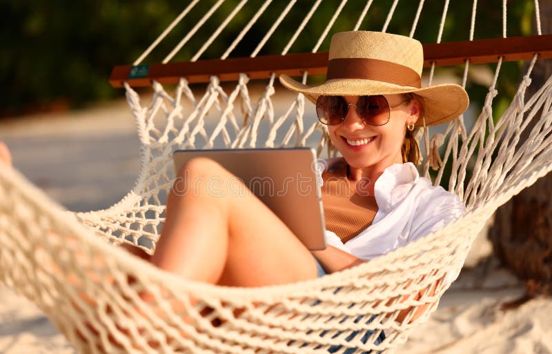Young smiling female, happy woman in straw hat and sunglasses using digital tablet while relaxing in the hammock on tropical sandy