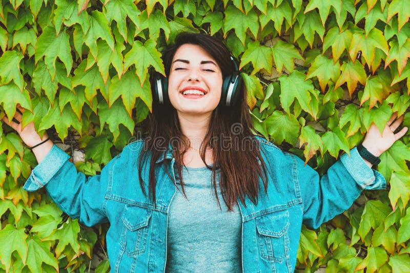 Young hipster girl listens and enjoys music leaning against an ivy wall - Pretty millennial woman relaxes with headphones in a