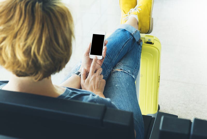 Young hipster girl sitting at airport in yellow boot on suitcase traveling in Europe, female hands texting message on gadget stock photography Young hipster girl sitting at airport in yellow boot on suitcase traveling in Europe, female hands texting message on gadget stock photography
