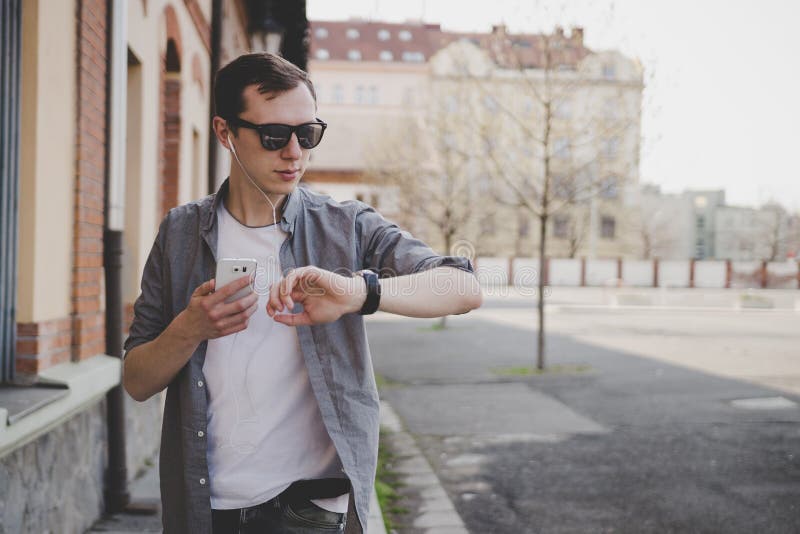 Young hipster man walking on the street, looking at watch and using his smartphone. With copy space.