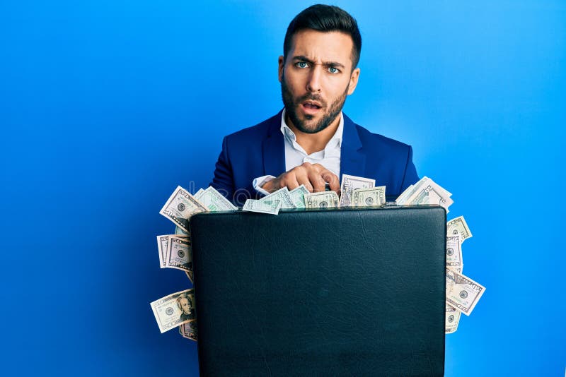 Young hispanic businessman holding briefcase full of dollars in shock face, looking skeptical and sarcastic, surprised with open