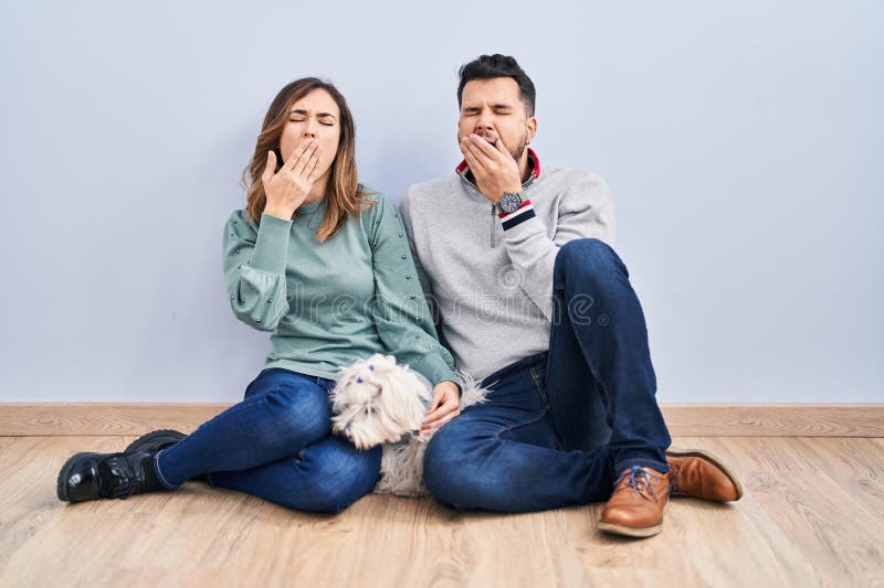 Young hispanic couple sitting on the floor with dog bored yawning tired covering mouth with hand