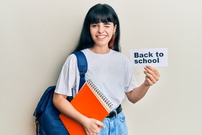 Young hispanic girl wearing backpack holding back to school banner winking looking at the camera with sexy expression, cheerful and happy face