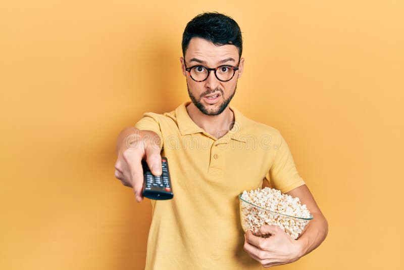 Young Hispanic Man Eating Popcorn Using Tv Control in Shock Face