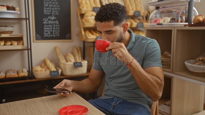 Young hispanic man enjoying coffee while using smartphone in a cozy bakery surrounded by bread and pastries stock image