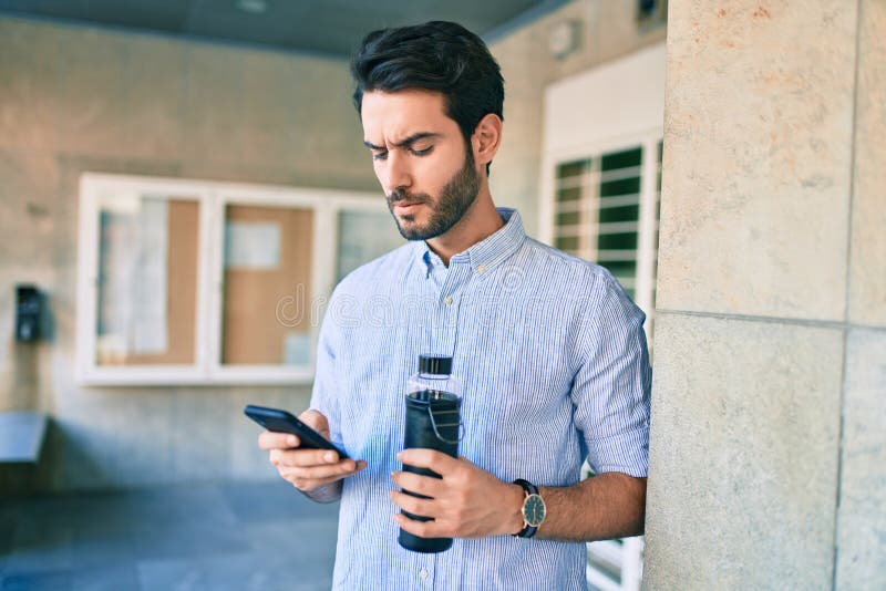 Young hispanic man with serious expression holding bottle of water and using smartphone at city stock images